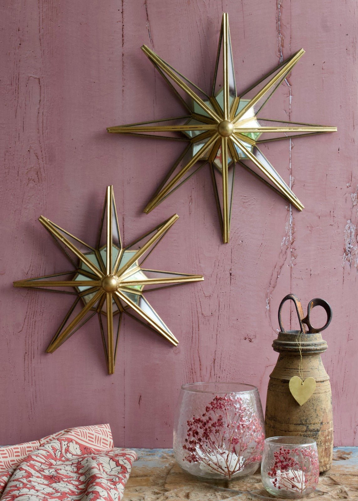 Two brass and mirror wall stars with a compact design, mounted on a pink wooden wall and styled with glass candle holders and rustic accessories.