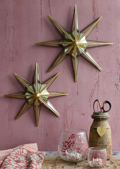 Two brass and mirror wall stars with a compact design, mounted on a pink wooden wall and styled with glass candle holders and rustic accessories.