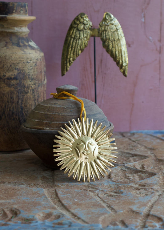 Brass sun ornament with facial detail resting on a rustic surface, with a clay pot and golden angel wings in the background.