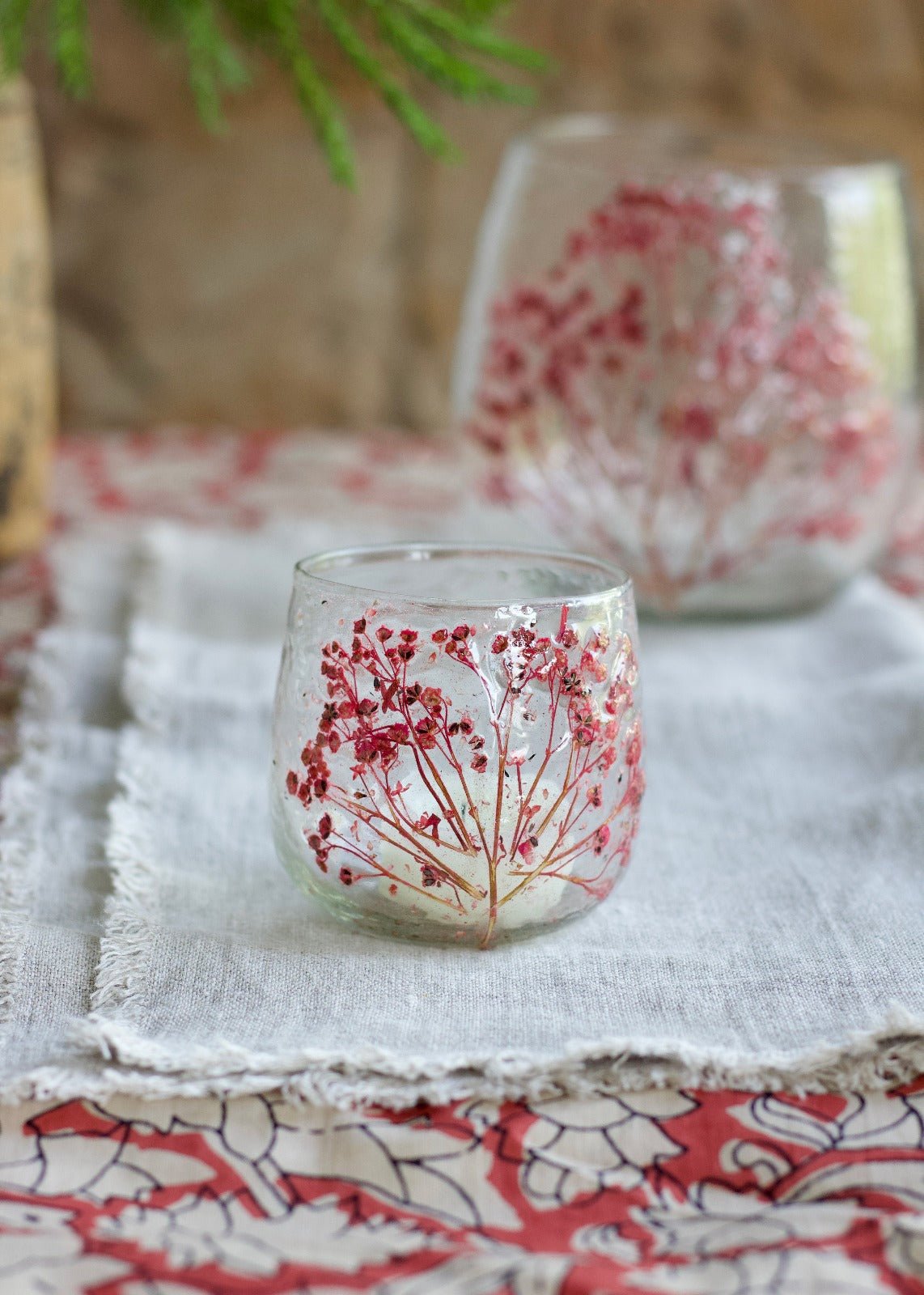 Handmade glass candle holder with dried red flowers, glowing with a tealight on a linen tablecloth.