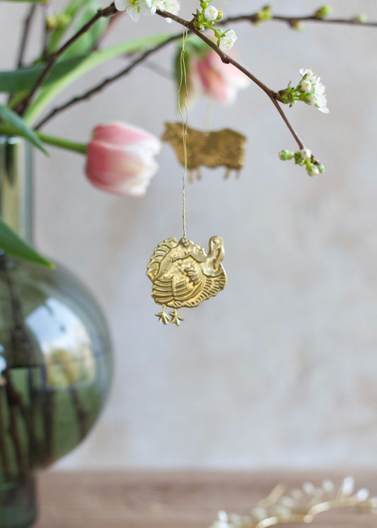 Brass turkey ornament hanging from a blossoming branch, with a tulip and green glass vase in the background.