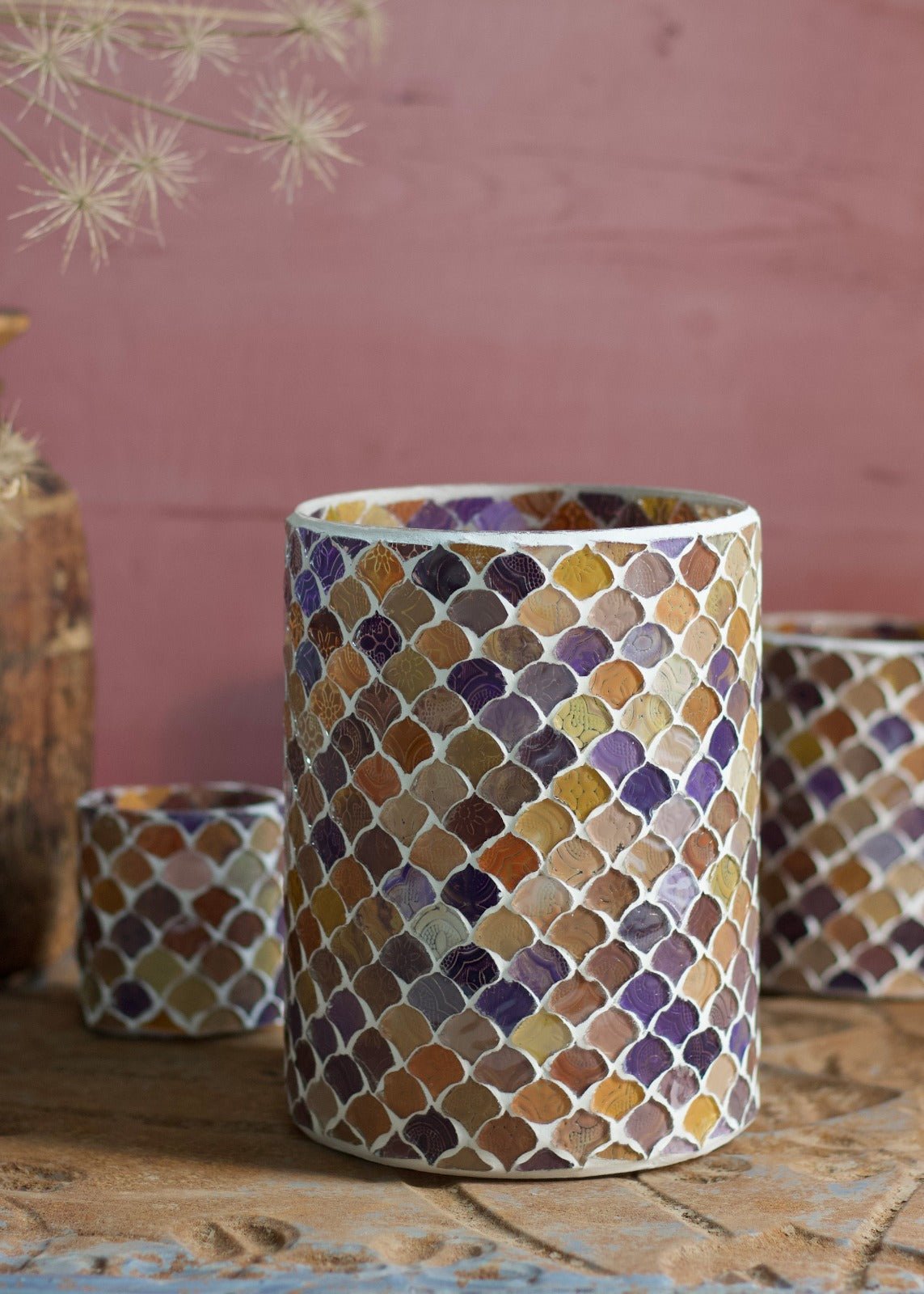 Large handmade mosaic vase in warm purple, ochre, and terracotta tones with a fish-scale pattern, styled on a carved wooden surface.

