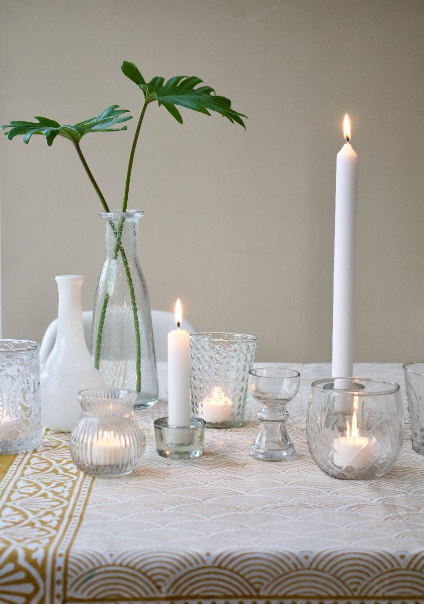 Small handmade clear glass vase with sculpted waist, styled on a dinner table with candles, other vases and green foliage.