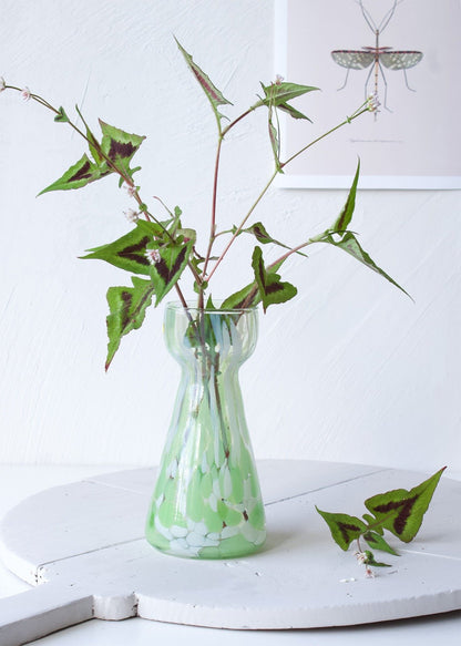 Handmade green and white speckled glass vase with hourglass shape, styled with wild foliage on a white table.

