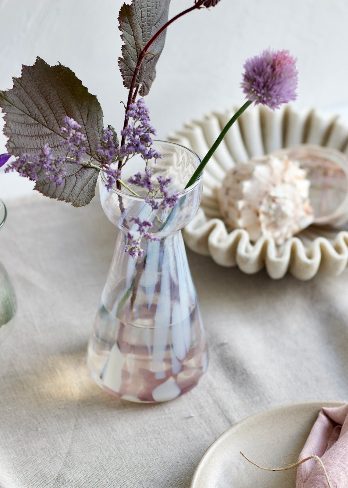 Handmade pink glass vase with white spots and narrow waist, styled with soft purple flowers and seashells on a linen table.