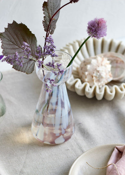 Handmade pink glass vase with white spots and narrow waist, styled with soft purple flowers and seashells on a linen table.