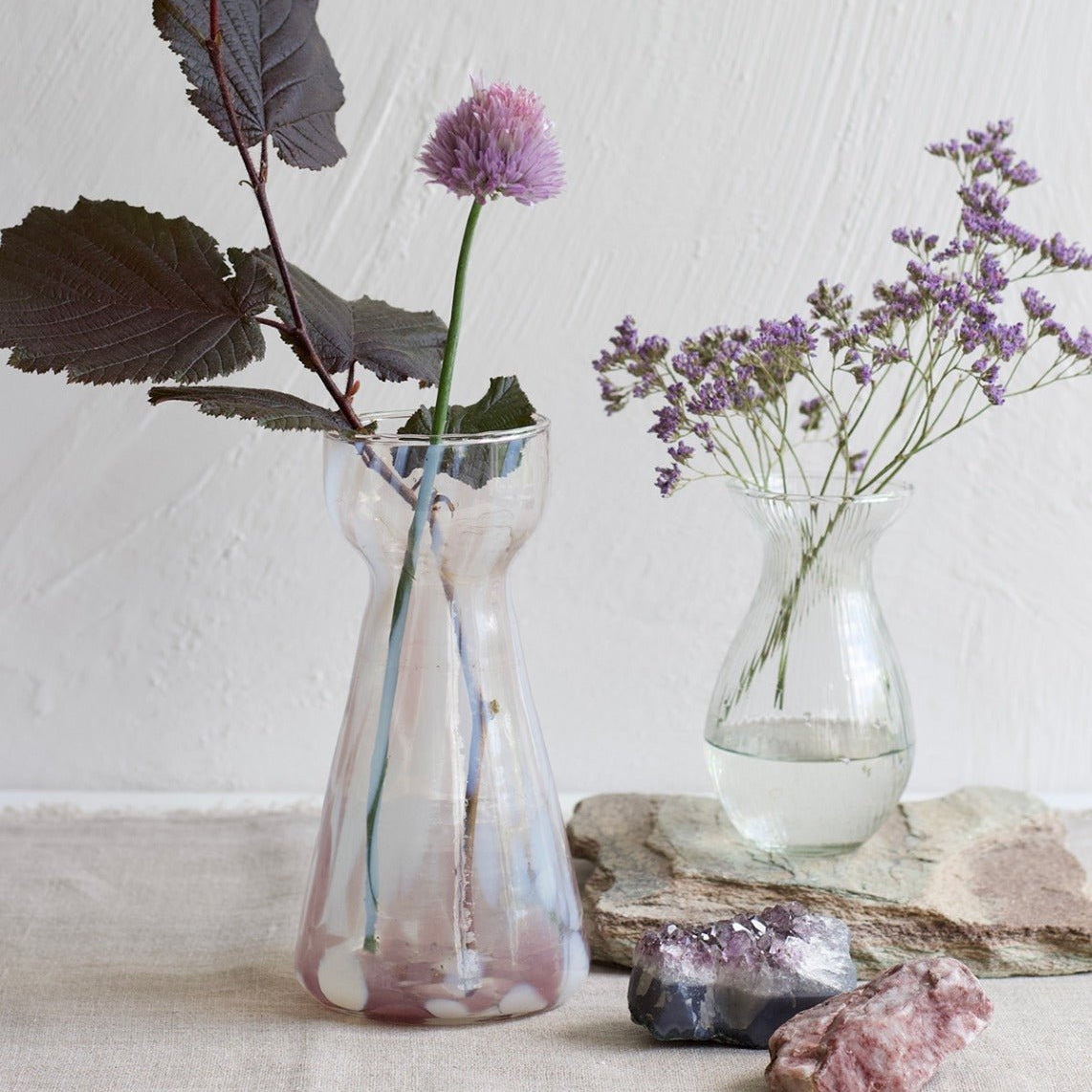Handmade pink and white glass vase with narrow waist, filled with purple flowers, styled next to a clear ribbed vase and gemstones on linen.

