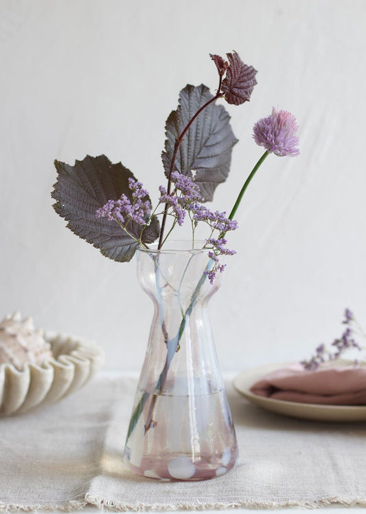 Handmade pink glass vase with soft white spots and hourglass shape, styled with purple flowers and dark foliage on a linen table.