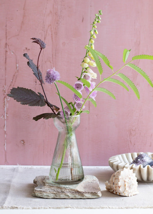 Tall ribbed glass vase with a narrow waist and flared top, styled with soft purple and green summer flowers, displayed against a dusty pink wall.