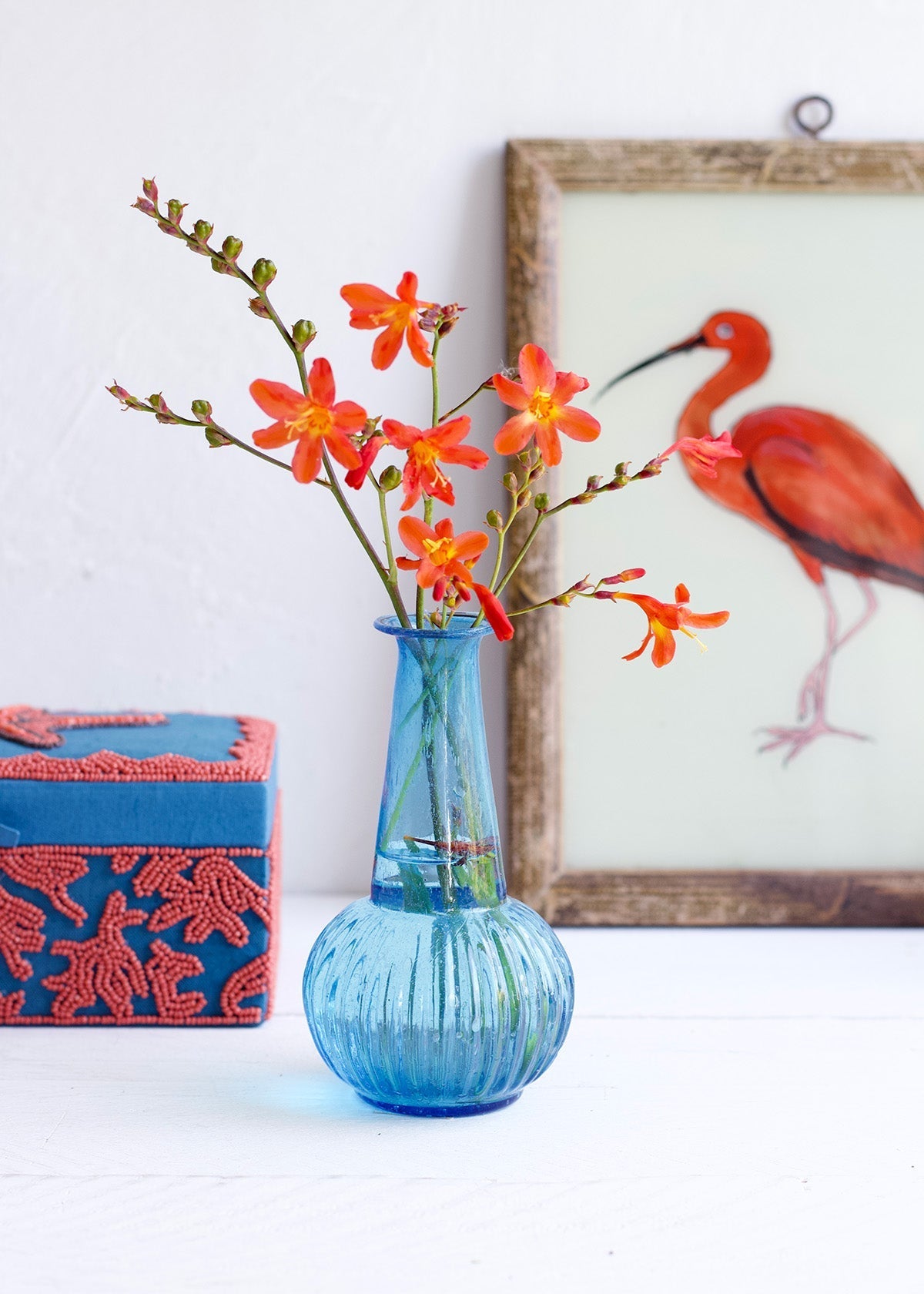 Blue vase with orange flowers on a white surface next to a red box and framed picture of a flamingo.