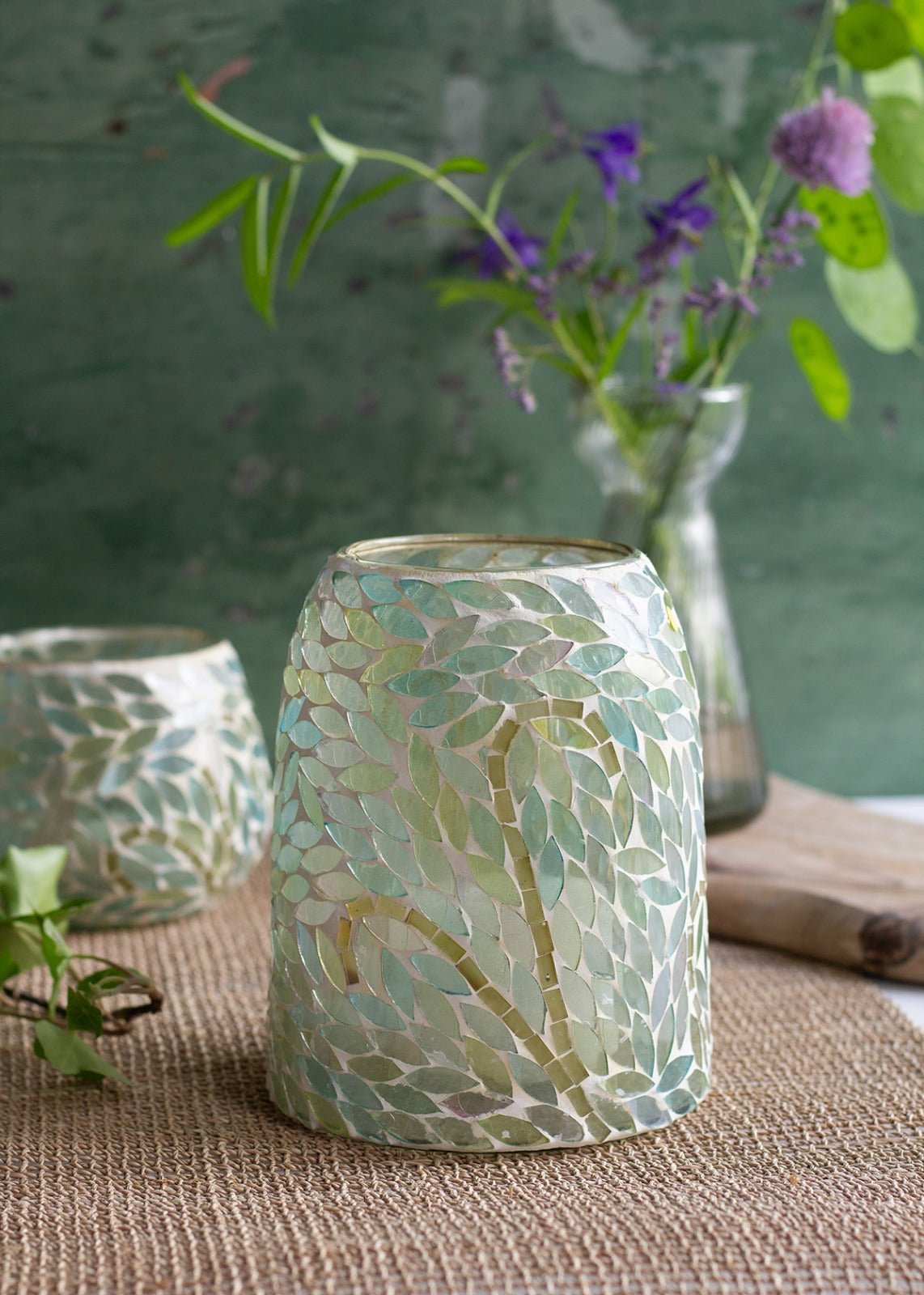 Small handmade mosaic glass holder in soft green leaf pattern, styled on a woven mat with wildflowers in the background.