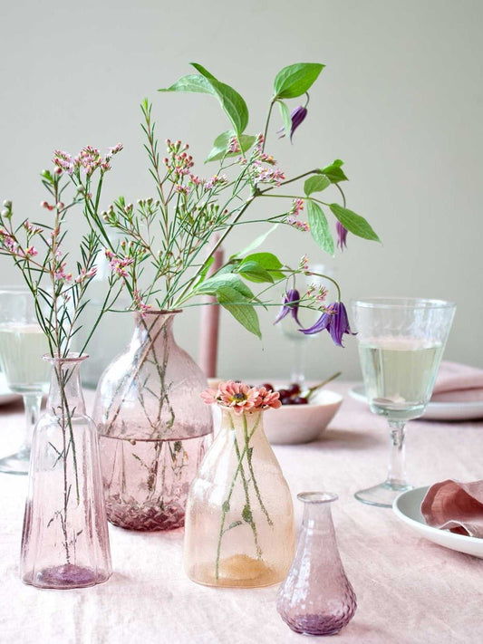 Delicate lilac and pink glass vases with wildflowers, styled on a pastel pink linen tablecloth with glassware and floral table decor.