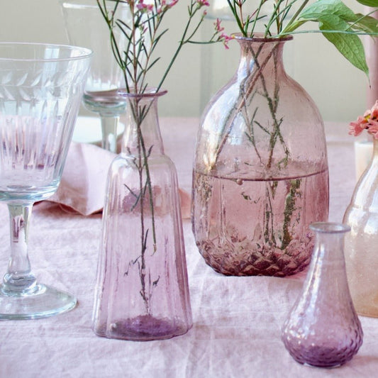 Assorted lilac and pink-tinted glass vases, hand-blown from recycled glass, styled with delicate wildflowers on a pastel table setting.