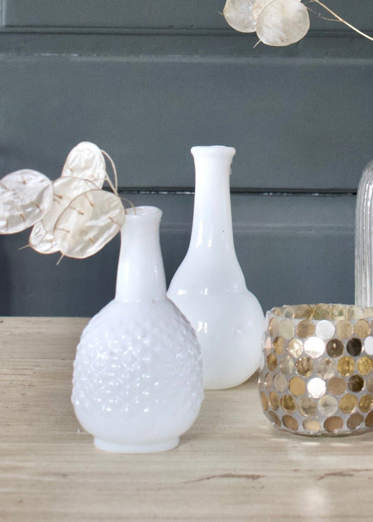 Two small white opaline glass vases on a wooden surface, one with a smooth surface and narrow neck, the other with a textured diamond pattern, styled with dried lunaria branches.


