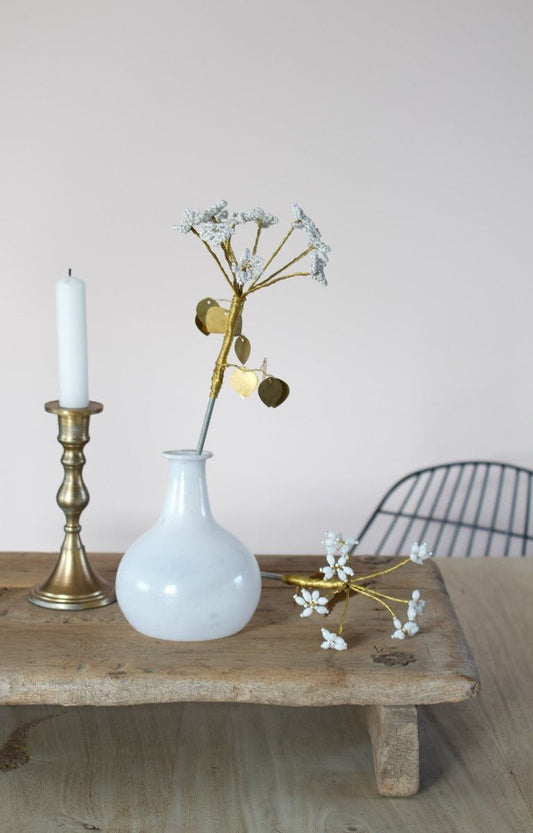White opaline glass vase with a rounded base and narrow neck, styled with a gold decorative flower and placed on a wooden platform next to a brass candle holder.


