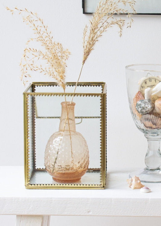 A small amber glass vase with dried grasses displayed inside a brass and glass display box on a white table, next to a tall glass filled with shells and vintage items.