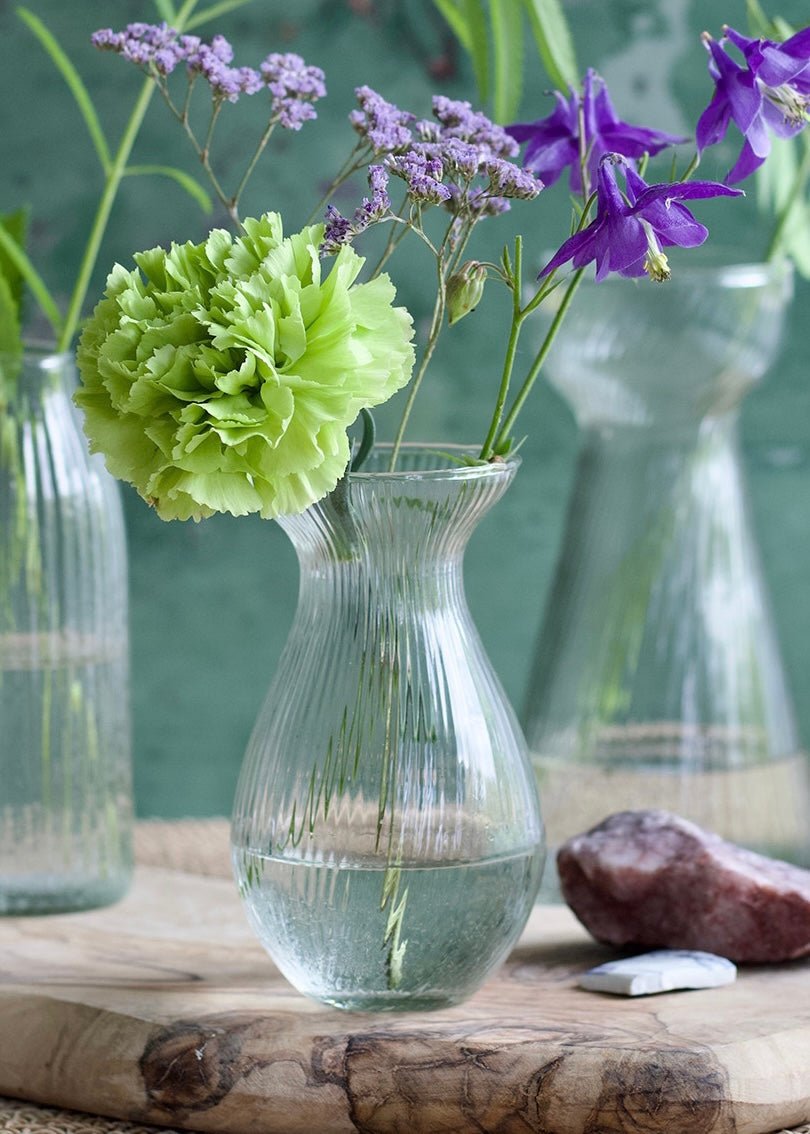 A pear-shaped ribbed glass vase filled with water and delicate spring flowers, including a vivid green carnation and purple statice. The vase sits on a wooden plank with natural stones, set against a rustic green background for a soft and organic look.