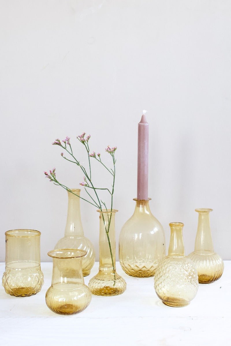 Collection of small hand-blown amber glass vases in assorted shapes, styled with a pink candle and delicate flowers on a white wooden surface.