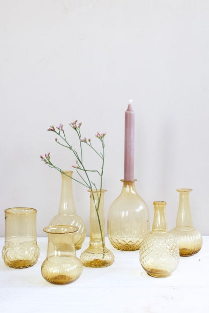 Collection of small hand-blown amber glass vases in assorted shapes, styled with a pink candle and delicate flowers on a white wooden surface.