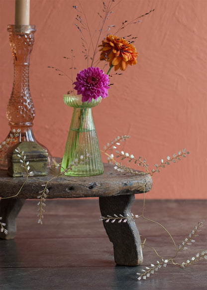 Decorative still life with a green vase, pink candle, and flowers on a wooden stool against a pinkish-brown background.