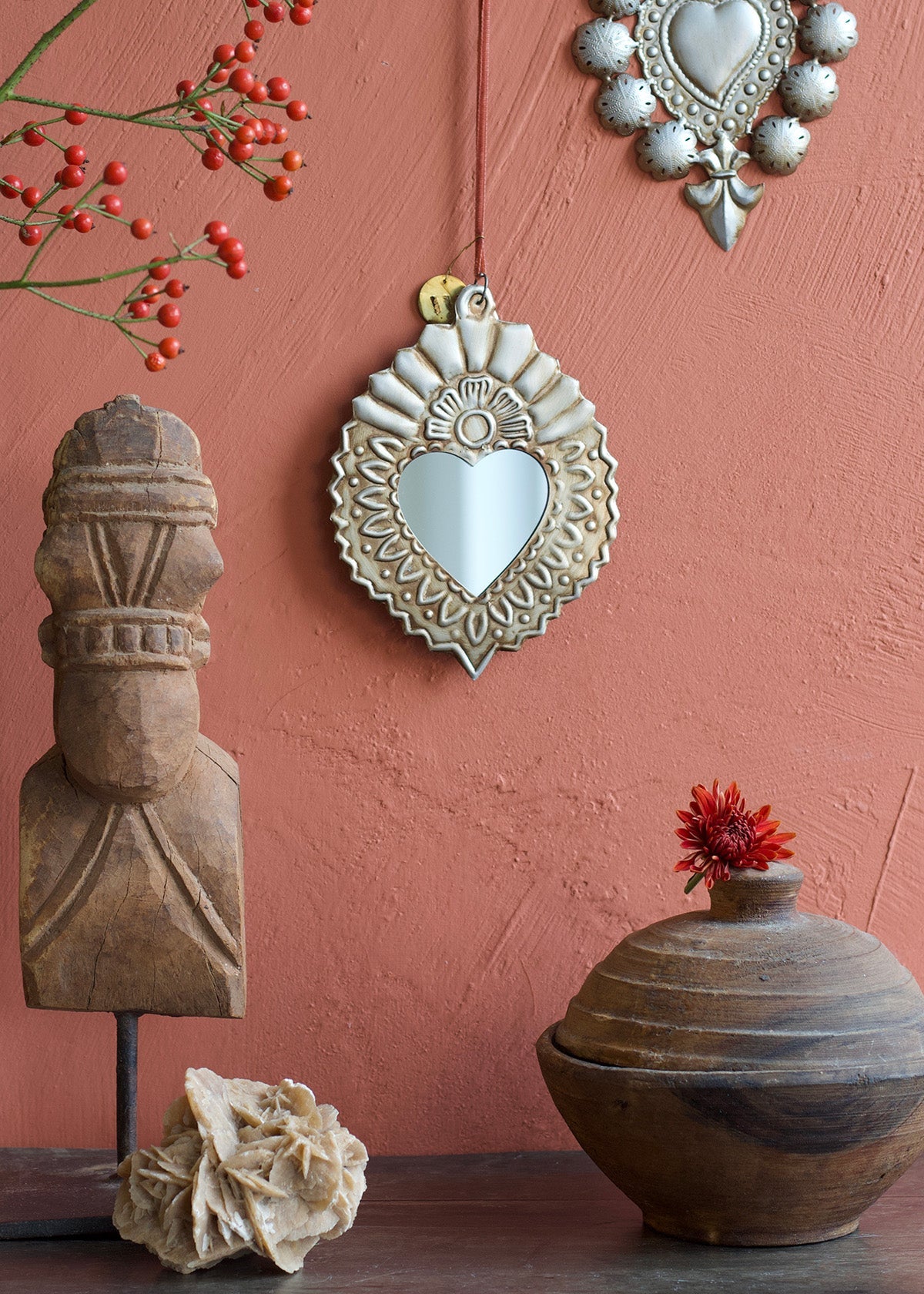 Decorative heart-shaped mirror with rustic sculptures and a vase against a pink wall.