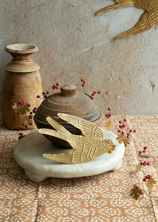 Brass swallow ornament resting on a marble stand, surrounded by red berries and rustic wooden pottery on a patterned fabric.