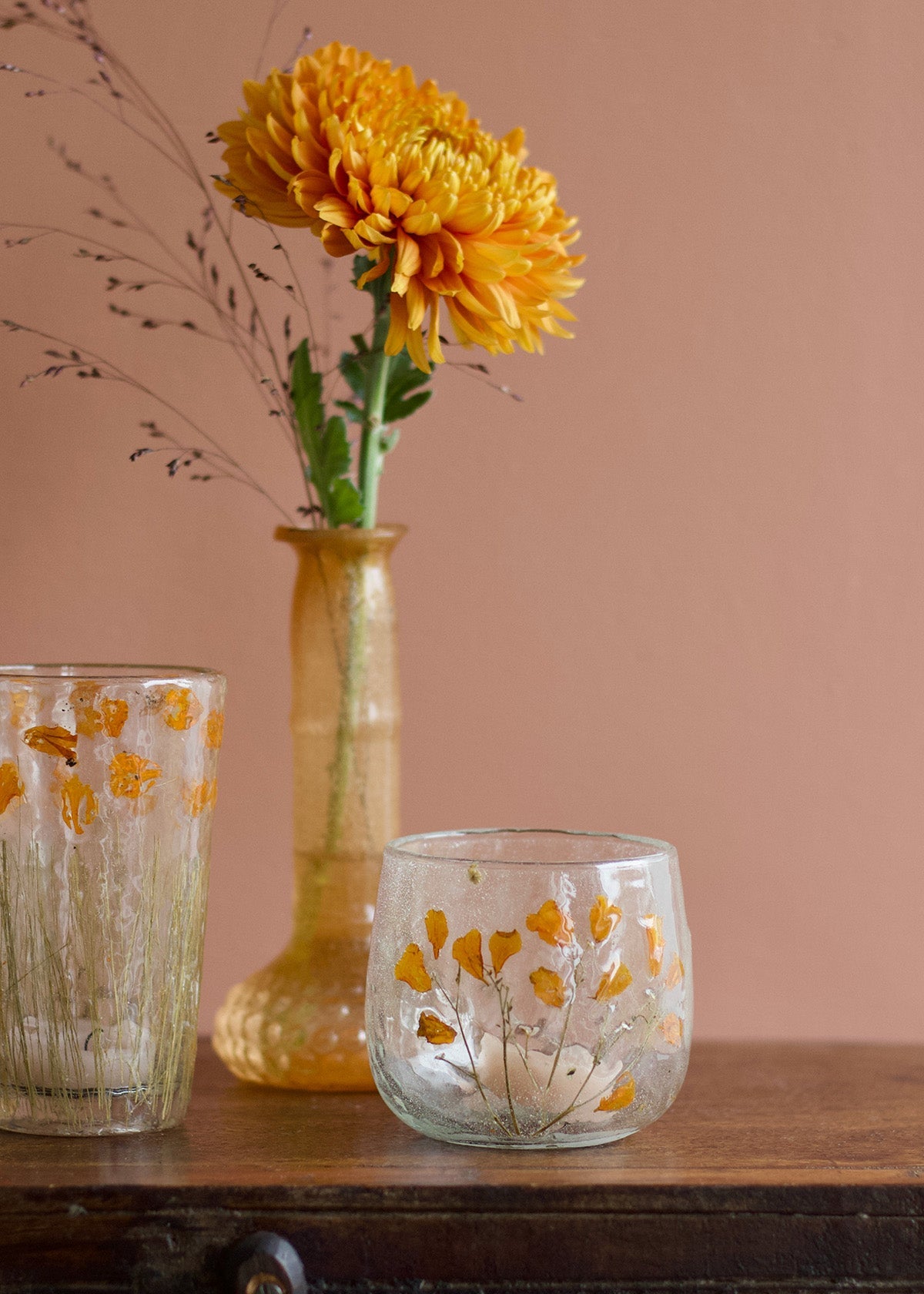 Two glass vases with yellow floral designs on a wooden surface against a pink wall.