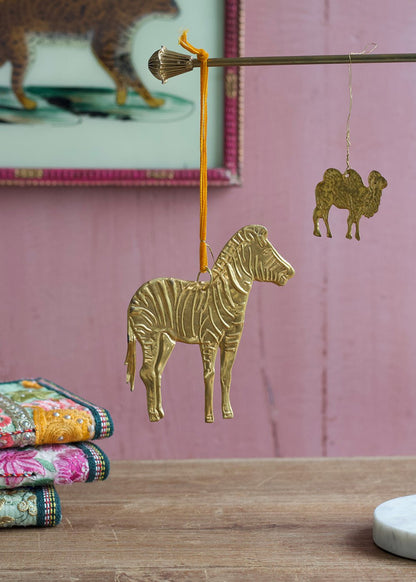 Brass zebra ornament hanging from a rod with velvet ribbon, accompanied by a small brass camel charm in the background, set against a soft pink wall.